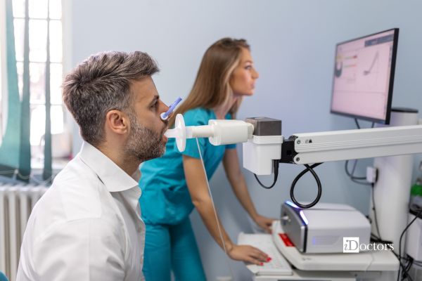 Paziente esegue una spirometria soffiando nel boccaglio dello spirometro, mentre un’operatrice sanitaria osserva i risultati sul monitor.
